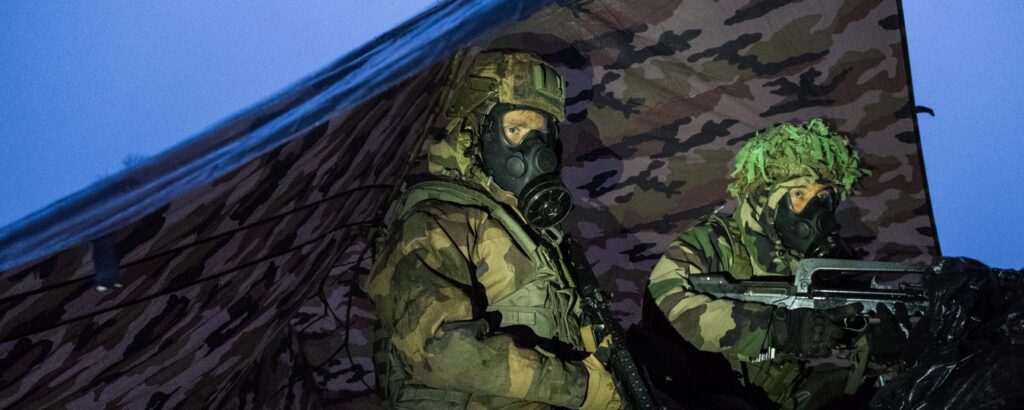 Two soldiers in camouflage and protective gear, including gas masks, using equipment developed by the CBD SBIR Program. They crouch under a camouflage tarp, one holding a weapon while the other observes the surroundings in a low-light environment, appearing alert.