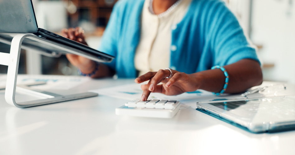 Accountant calculating firm-fixed-price contract costs for a government contractor using a calculator and laptop at a desk.