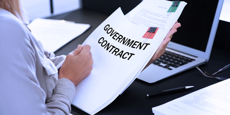 Person reviewing a document labeled ‘government contract’ at a desk with a laptop, illustrating FAR and CAS guidelines for government contractors.